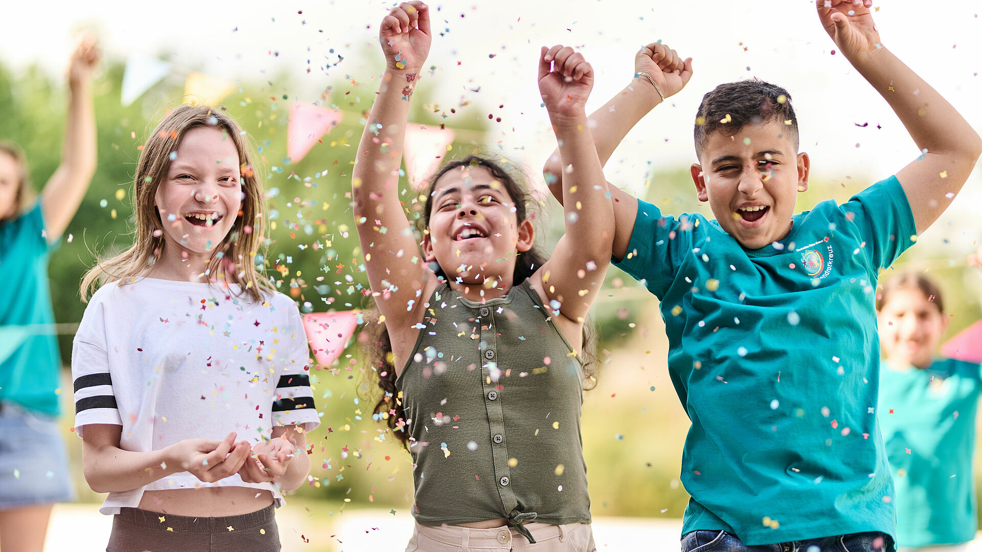 Zu sehen sind drei Kinder, die freudig in die Luft springen und Konfetti werfen. Eins der Kinder hat ein JRK-T-Shirt an.