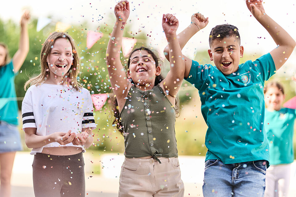 Zu sehen sind drei Kinder, die freudig in die Luft springen und Konfetti werfen. Eins der Kinder hat ein JRK-T-Shirt an.