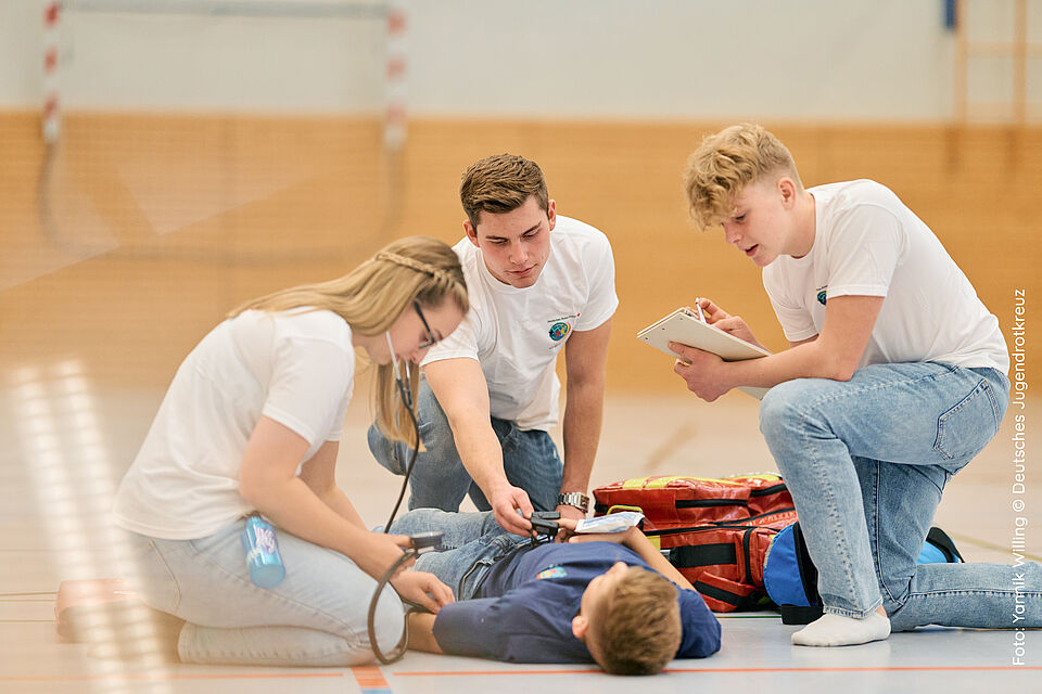 Zu sehen sind vier Personen in einer Sporthalle. Eine liegt auf dem Boden, die anderen drei knieen um sie herum. Diese drei Personen sind Schulsanitäter:innen und kümmern sich um die Person, die auf dem Boden liegt.